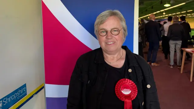 Rosemary is wearing a black top with a red Labour party rosette. She has short grey hair and wears round dark glasses.