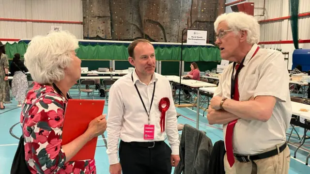 Two men wearing white shirts and a woman wearing a white dress with poppies on it at the count, which is being held inside a sports hall. They are members of the Labour party who are in discussion.