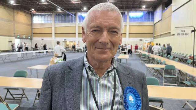 Phil Bibby a man with grey/white hair is standing in a hall with tables and chairs. He is wearing a grey suit jacket and green and blue striped shirt and is looking directly towards the camera.