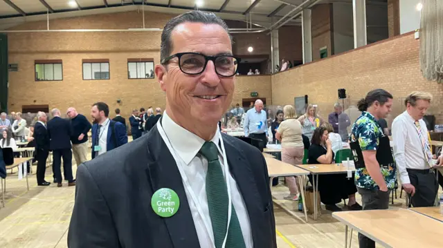 Ben Ensch wearing a black suit and a Green Party badge. He is standing in a sports hall where the election count is taking place. Tables and lots of people behind him.