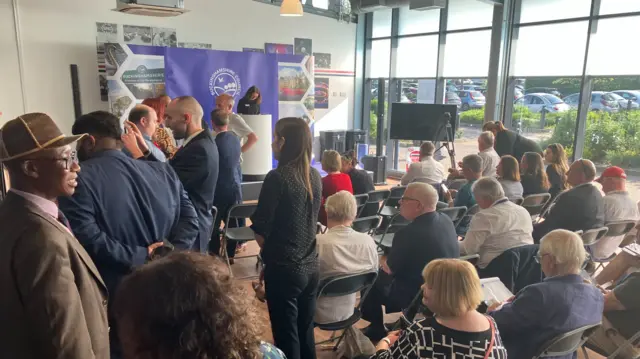 People fill a room at Stoke Mandeville Stadium including people on rows of chairs in front of a platform.