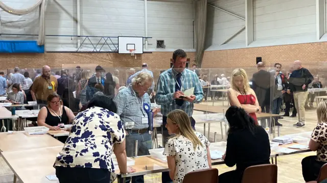 Ryan wearing a blue checked shirt standing behind a glassed barrier taking notes