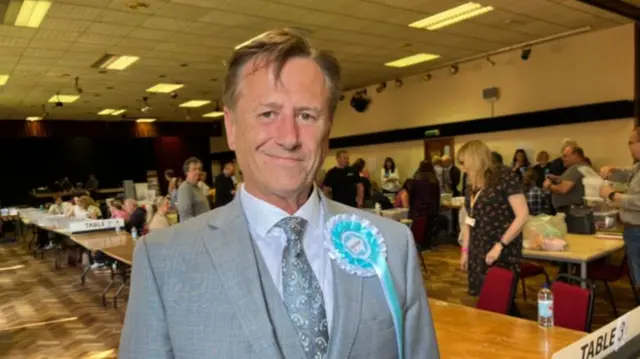 A man with brown hair a grey suit and a large blue rosette in a large room