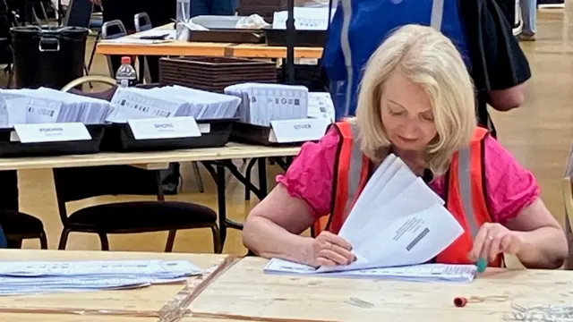 A blonde woman in a pink shirt and orange vest flips through ballot papers