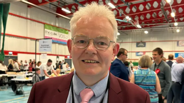 Ian McCord wearing a burgundy suit jacket, blue shirt and pink tie. He is smiling at the election count.