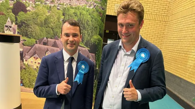 Lewis Cocking (left) and Alexander Curtis standing in front of a raised platform in a sports hall. Cocking is wearing a blue suit and tie. Curtis is wearing a blue jacket and light brown/cream trousers and no tie. Both are giving a thumbs-up.