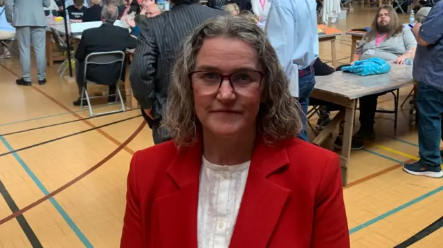 A woman in a sports hall wearing a red blazer and white top