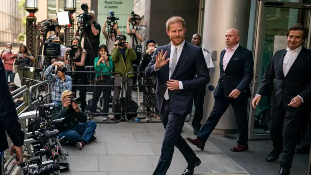Prince Harry exits the Rolls Buildings in London. A number of members of the press are outside holding cameras to take photos.