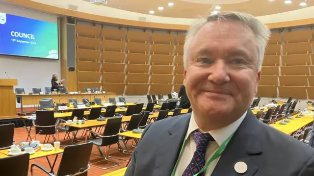 A picture of independent councillor Stuart Wilson in the council chamber in Aylesbury. He has grey/white hair, and is wearing a dark jacket and a tie that is blue but with white lining and red flowers on it