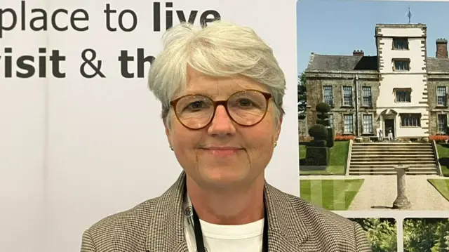 Anna Earnshaw has short grey hair and is wearing glasses, a chequered jacket and a white T-shirt. She is standing in front of a large banner which has a grand country house on it.