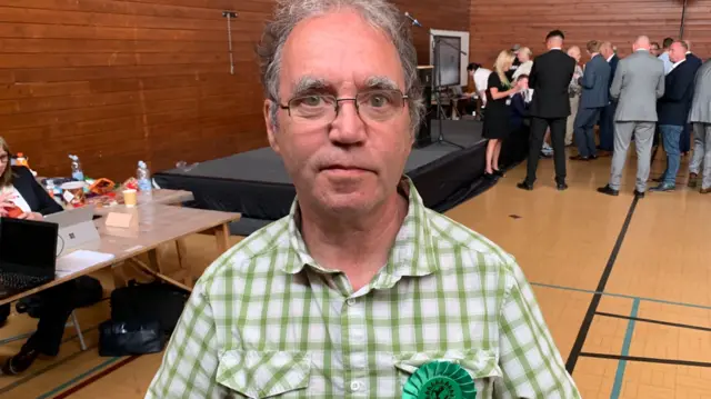 A man in a green check shirt, she is standing in a sports hall and wearing a green rosette.