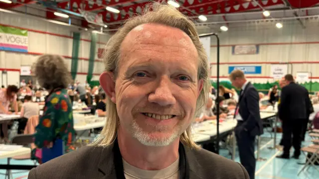 Jonathan Harris with his long, fair hair brushed back. he is wearing a brown suit jacket over a grey T-shirt while standing inside the sports hall. People are counting votes behind him.