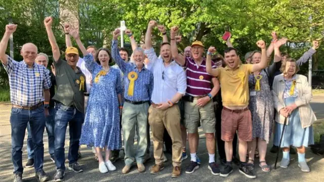 A group of people photographed outside. They have their hands in the air and are cheering. Some are wearing yellow Liberal Democrats rosettes.