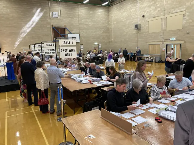 Dozens of people sit at tables in what looks like a sports hall, counting papers.