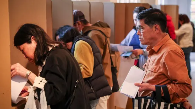 People queue at voting booths with folded pieces of paper in their hands