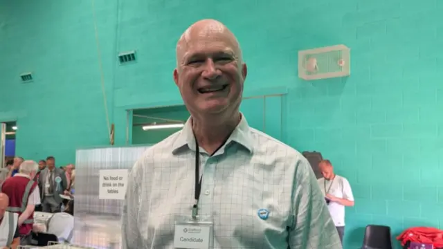 A man with a bald head and a lanyard in a room with turquoise walls