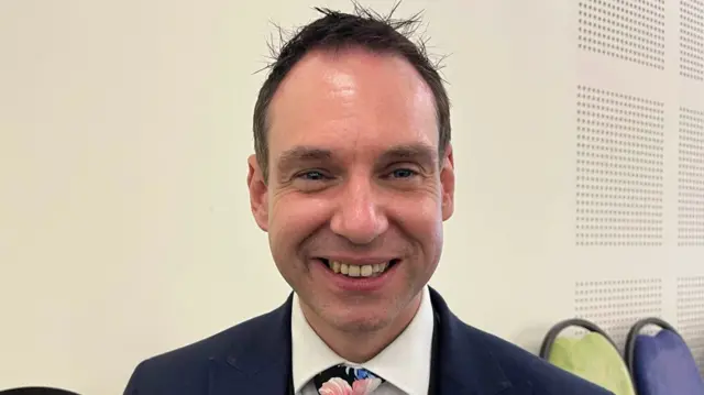 Daniel Lister with short brown hair smiling and wearing a suit.