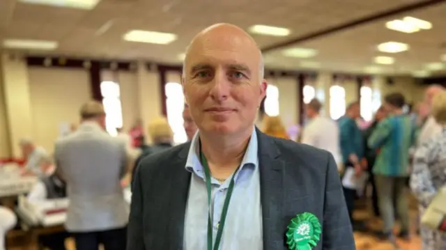 A man with short grey hair, a pale blue shirt and dark jacket with a green rosette in a large room