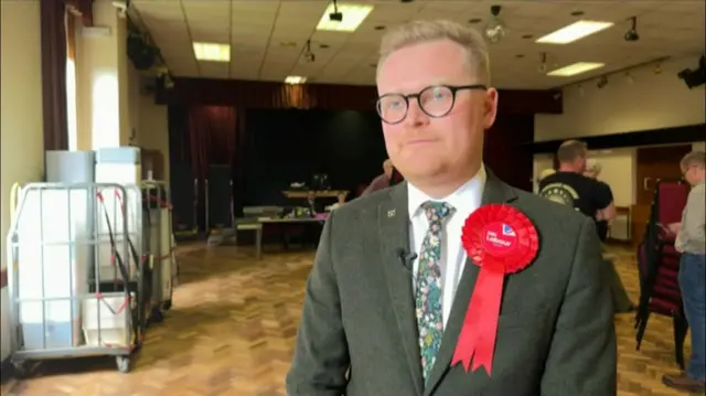 A man with short blonde hair, a grey suit and a large red rosette in a large room with a wooden floor