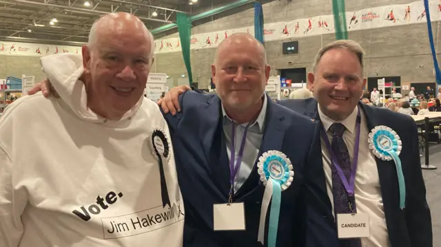Jim Hakewill, Christopher Kellett and Brian Bennyworth all standing with their hands around each others' shoulders. Hakewill has a white hoodie on, while the others are wearing suits. All are smiling.