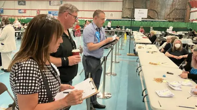 Two men and a woman watch on with clipboards as votes are counted by people sat behind a long table.