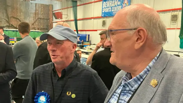 Ray Connolly wearing a black shirt and grey cap. He is standing next to an older man in the sports hall.