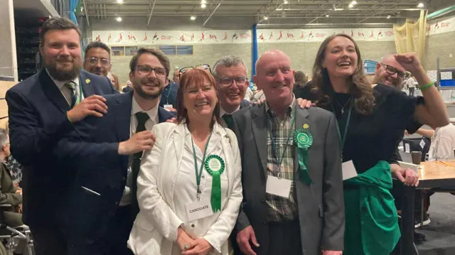 Green Party members celebrate together in a huddle after their latest victory. They are all smartly dressed with different green features on their outfits, including lanyards, ties and rosettes.