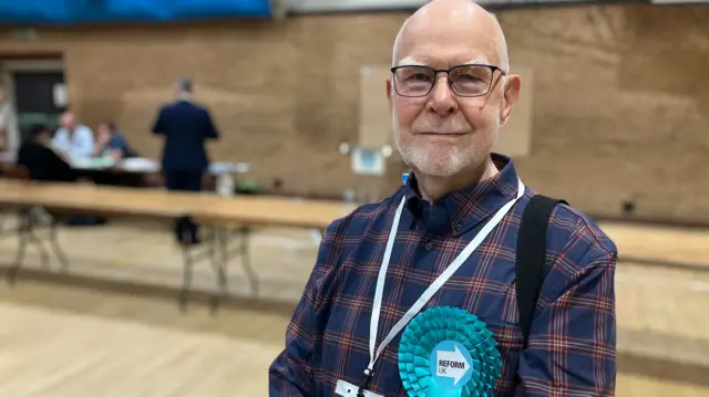 John Roberts wearing a dark blue checked shirt. He has a white lanyard hung around his neck and a light blue Reform UK rosette attached to his shirt.