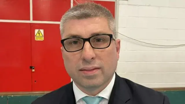 Mark Arnull wearing a pale blue tie, white shirt, black suit jacket and black glasses. He has short grey hair and is sitting in a sports hall.
