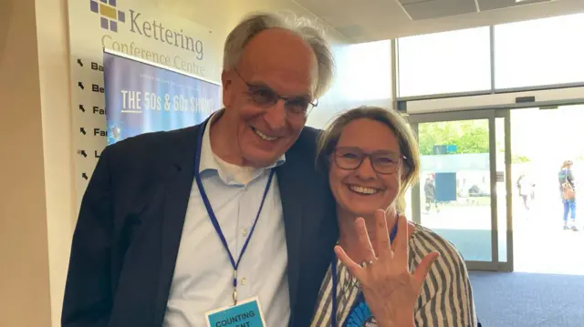 Peter Bone wearing a black suit jacket and pale blue shirt. He is leaning on Helen Harrison, who is holding her hand up to show her engagement ring. They are both smiling.