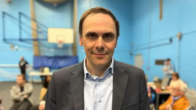 A man with short dark hair, a white shirt and a dark grey jacket in a room with blue walls
