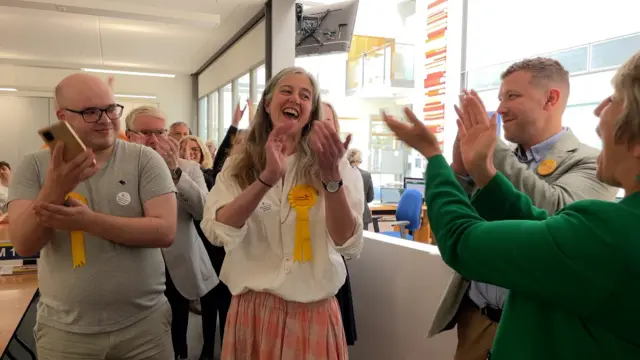 A group of people, each wearing yellow Liberal Democrat rosettes are clapping and celebrating with their hands in the air