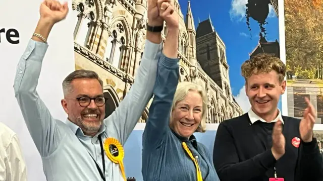 The successful Lib Dem candidates hold their hands in the air in celebration on stage. Another candidate smiles and claps.