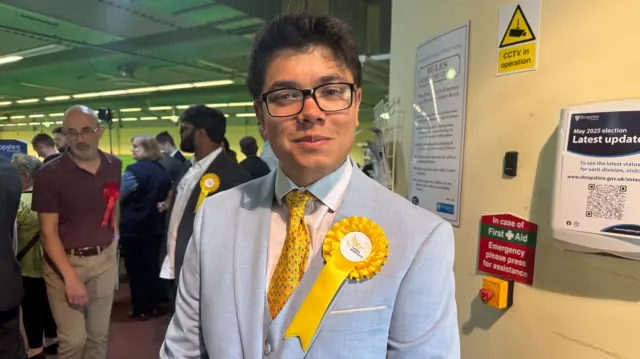 Gregory Ebbs is wearing a baby blue suit with a yellow tie and yellow lib dem rosette badge