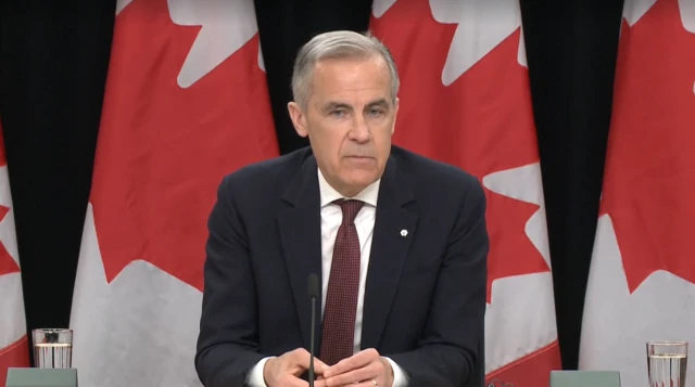 Carney headshot as he sits at table in front of Canada flags