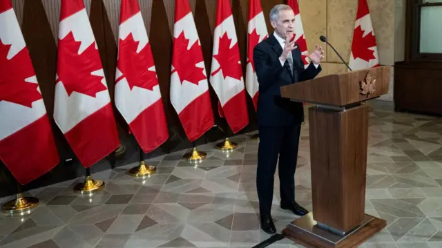 Mark Carney speaks at a podium embossed with a carved maple leaf at the front. He is wearing a black suit and trousers over a white shirt and black shoes. Behind him is an arrangement of Canada's red and white flags on a stand.
