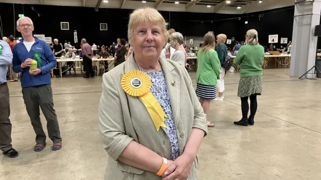 A woman in a hall where local election votes are being counted. She is wearing a yellow rosette