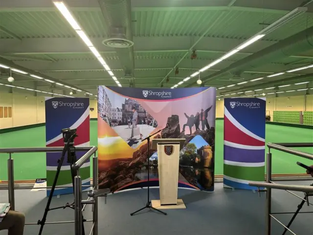 An empty wooden podium in front of a Shropshire council banner. A bowling green is in the background