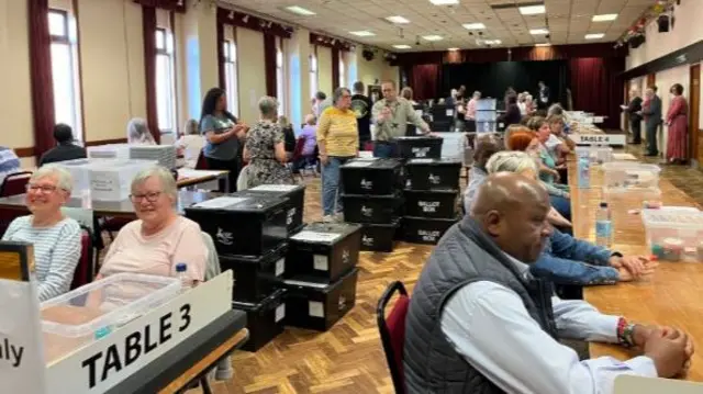People sat at rows of tables with boxes and papers on them