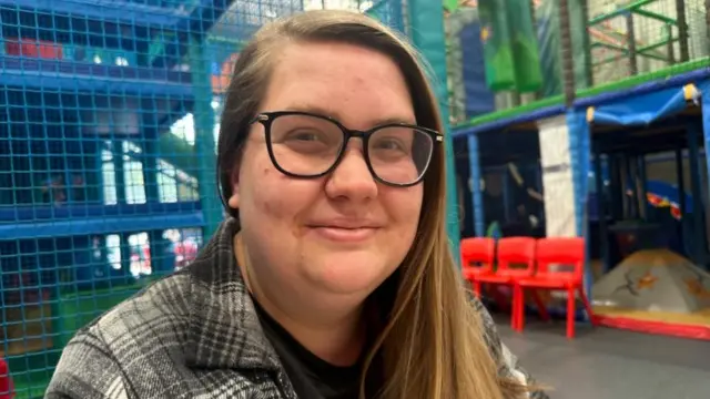 A woman sitting at a table in soft play centre. She is wearing a grey plaid shirt and square glasses. She is smiling.