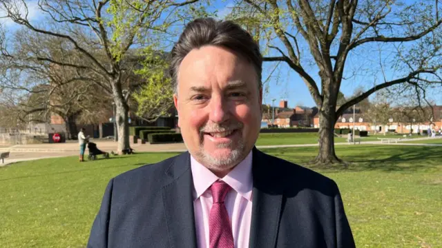 A man standing in a park. He is wearing a pink shirt and tie and a navy suit.