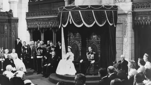 royals sitting at a throne surrounded by people. It's in black and white