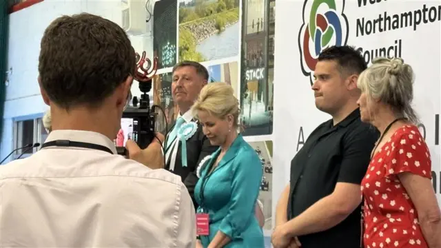 Four candidates stand on stage as the results are read out. The back of a man holding a camera blocks part of the image to the left.