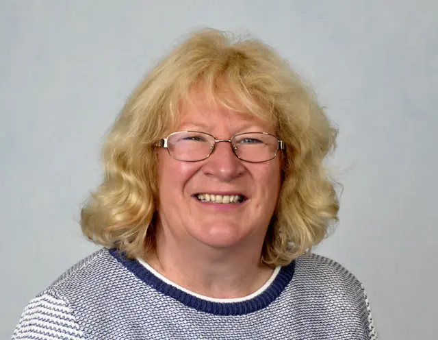Woman with blonde hair and wire-framed glasses, wearing a blue and white jumper.