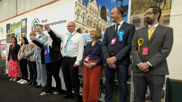 Reform UK councillors on stage pump the air together in celebration. They are flanked by other candidates who smile and watch on.
