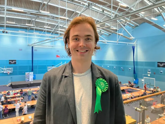 Peter Charles Rees smiles at the camera inside a counting centre. He is wearing a dark jacket over a grey T-shirt, with is wearing a Green Party rosette.