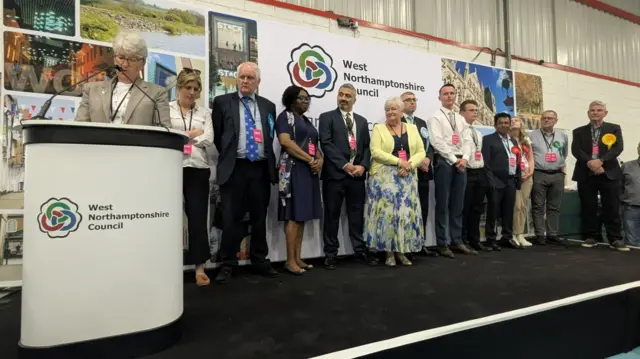 A large group on candidates lined up on stage as the council chief executive reads the results from behind a lectern.
