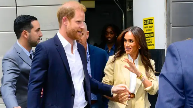 Harry and Meghan leaving an event, several people in suits surround them.