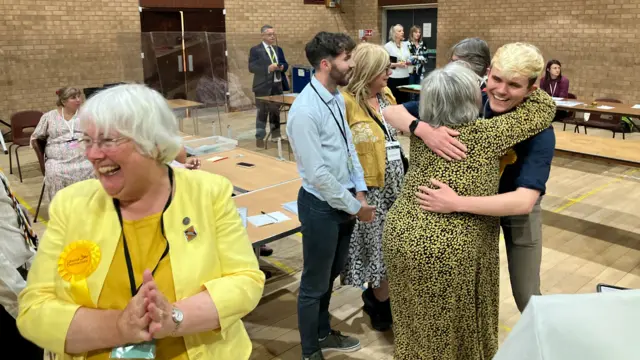 Tom wearing a blue shirt hugging Lorna Dupre, who is wearing a yellow and black dress