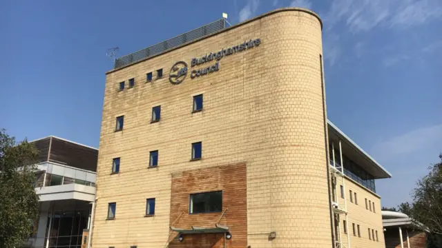 A picture of Buckinghamshire Council's headquarters in Aylesbury, which is a beige building of five floors. There is blue sky behind the picture.